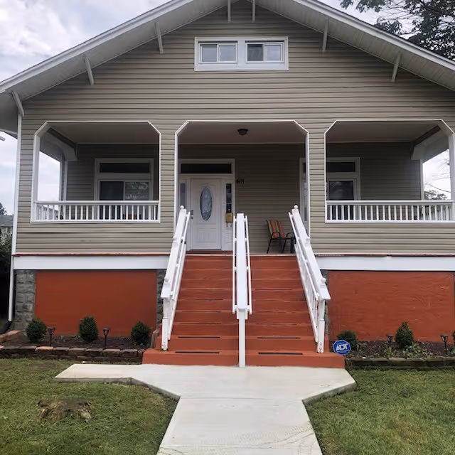 Front exterior view of a single-story house with a covered porch, white railings, and a central staircase leading up to a white front door with an oval glass panel. The house has beige siding, a red foundation, and small shrubs planted along the front.