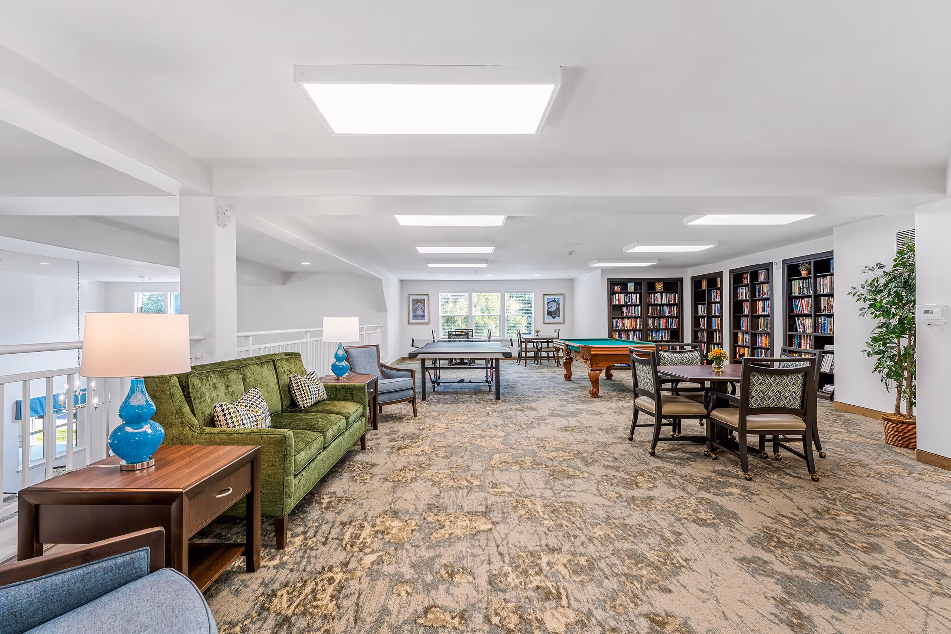 A spacious, well-lit common area with a green sofa, blue lamps on wooden side tables, several chairs, a ping pong table, a pool table, and bookshelves filled with books along the far wall. There are also tables and chairs for sitting and socializing, with large windows letting in natural light.