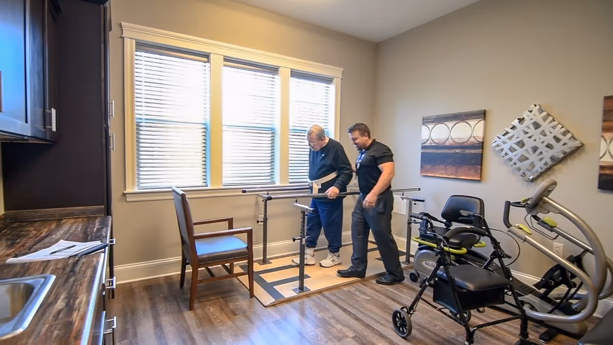 An elderly man is assisted by a caregiver while using parallel bars for walking practice in a room with wooden flooring, a chair, exercise equipment, and large windows with blinds.