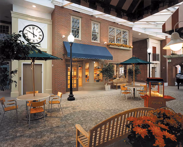 Indoor common area designed to look like an outdoor street scene with brick walls, large clock, street lamps, tables with green umbrellas, chairs, benches, a popcorn machine, and potted plants.