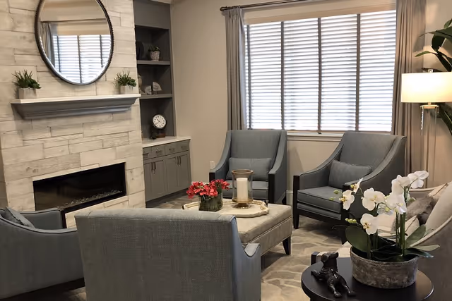 A cozy living room with four blue upholstered armchairs arranged around a square ottoman with a candle and flowers on top. There is a modern fireplace with a round mirror above it, built-in shelves with decorative items, a large window with blinds and curtains, a floor lamp, and a small table with a white orchid plant and a decorative figurine.