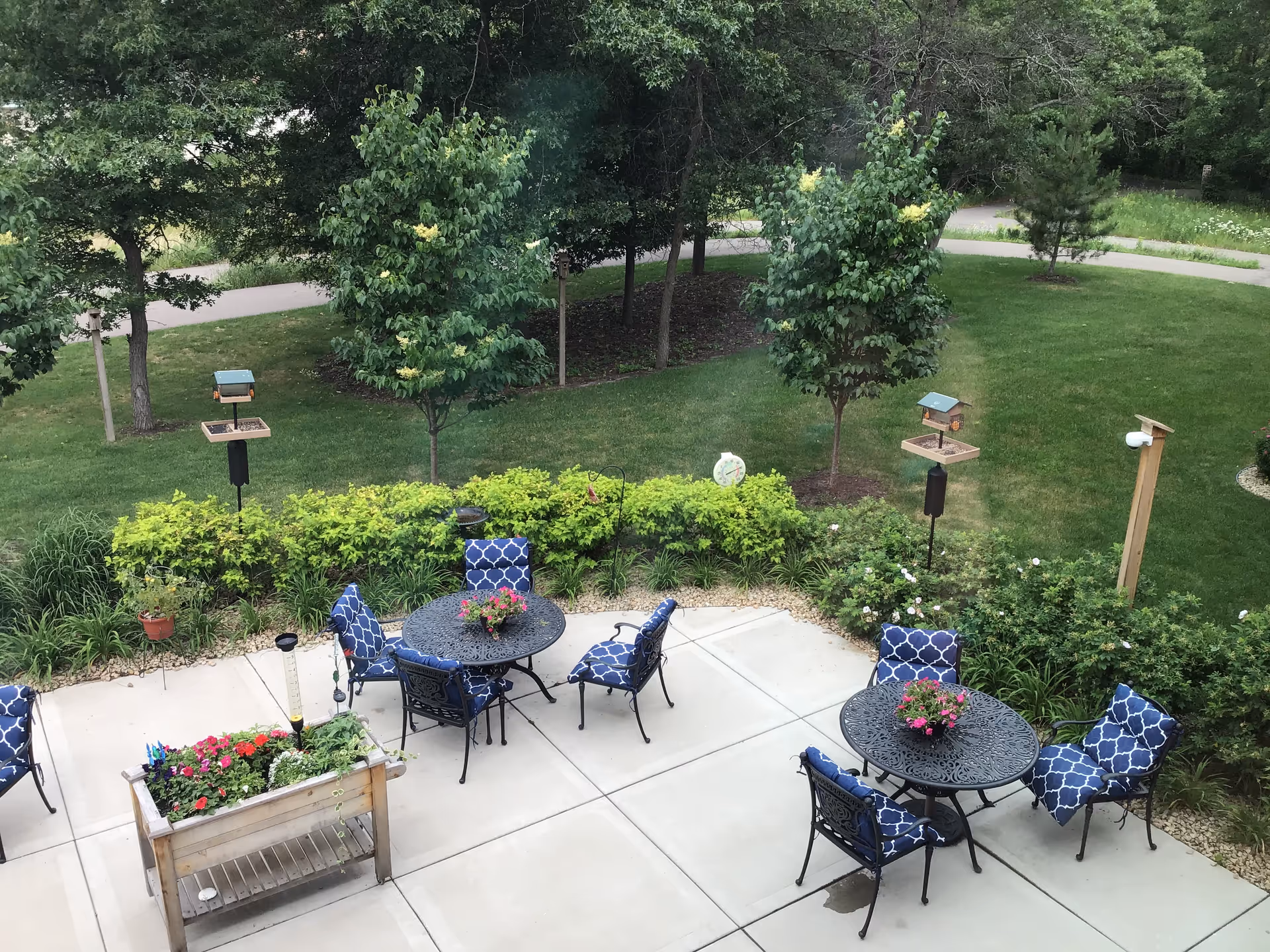 Patio with round metal tables and blue-patterned cushioned chairs overlooking a grassy yard with trees and bird feeders.