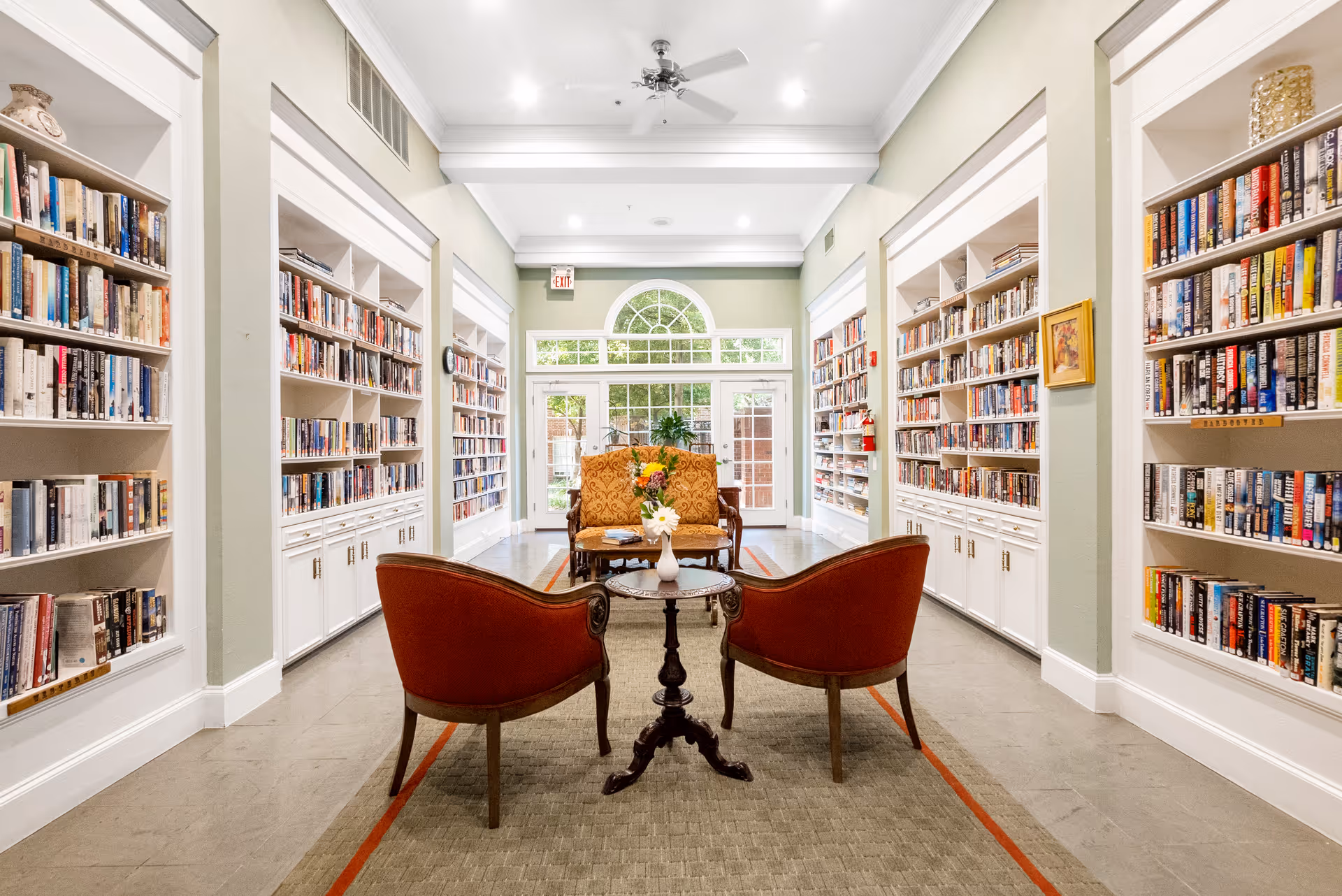 A bright and spacious library room with tall white bookshelves filled with books lining both sides. In the center, there are two red upholstered chairs facing a small round wooden table with a vase of flowers. Behind the table is a patterned loveseat. Large windows and a glass door at the far end let in natural light, with a view of greenery outside. The room has a high ceiling with recessed lighting and a ceiling fan.