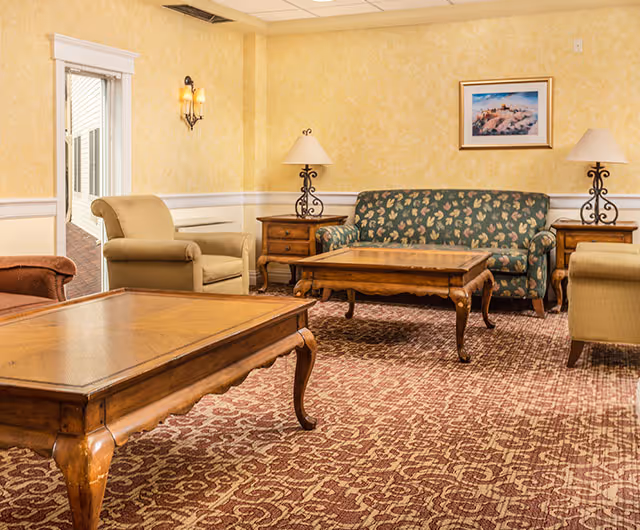 A cozy living room with patterned carpet, a floral upholstered sofa, two beige armchairs, two wooden coffee tables, two wooden side tables each with a lamp, yellow textured walls, and a framed picture hanging on the wall.