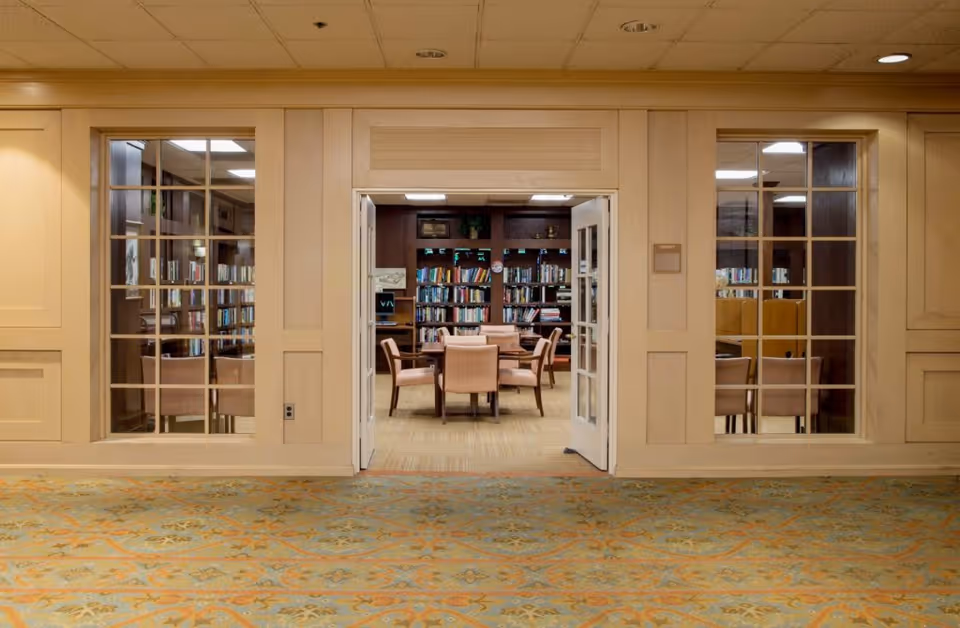 View of a library room through open double doors in a senior living facility. The room contains a round table with six upholstered chairs and bookshelves filled with books along the back wall. The walls around the doorway have large windows with wooden frames, and the floor outside the room is carpeted with a patterned design.