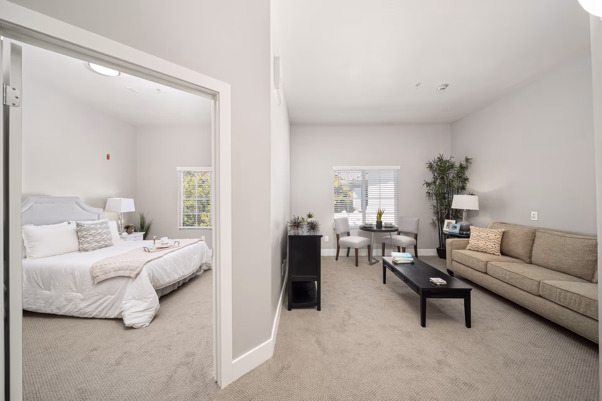 Bright apartment-style living area with a sofa, coffee table and small dining nook, with a bedroom visible through an open doorway.