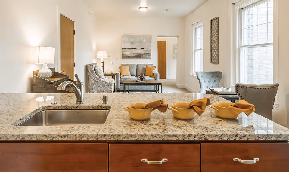 View of a modern living room from a kitchen island with a granite countertop and a stainless steel sink. The living room features a gray sofa with orange cushions, two patterned armchairs, a coffee table, and large windows letting in natural light. A lamp and wall art are also visible.