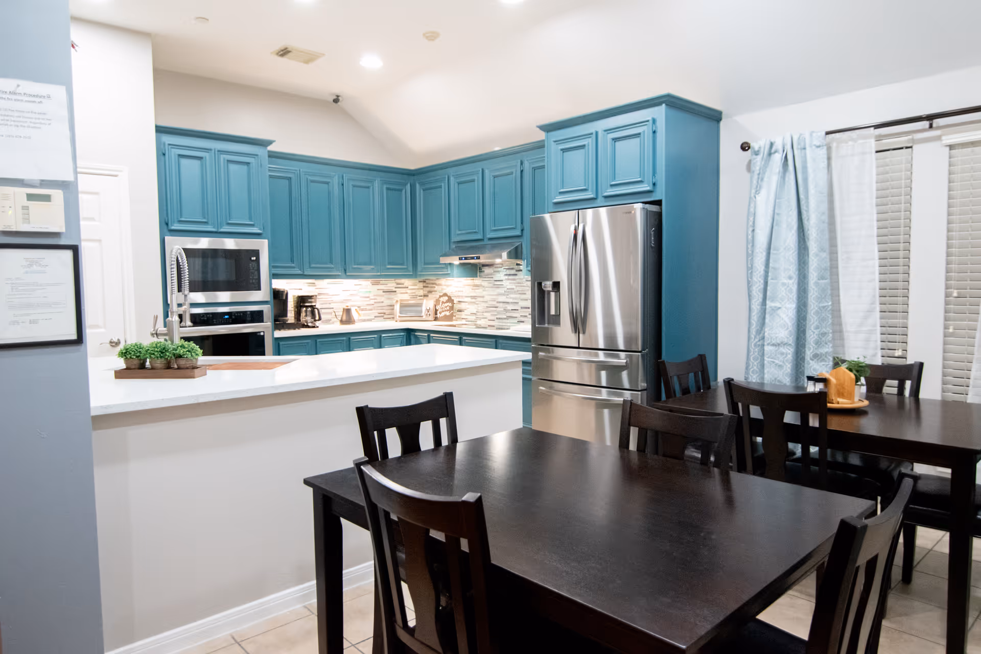 Communal dining area with dark wood tables and chairs adjacent to a modern kitchen with teal cabinets and stainless-steel appliances.
