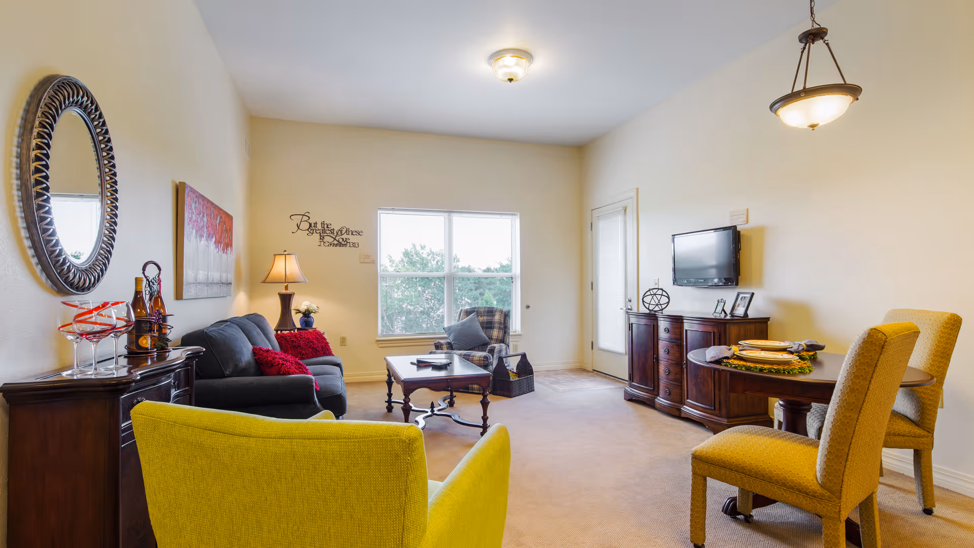 A cozy living room with a yellow armchair in the foreground, a dark gray sofa with red cushions, a wooden coffee table, and a plaid armchair near a window. There is a round dining table with two yellow chairs on the right side, a wall-mounted TV above a wooden cabinet, and a decorative mirror and lamp on a sideboard on the left wall. The room has beige walls and carpet, with a ceiling light and a hanging light fixture above the dining table.