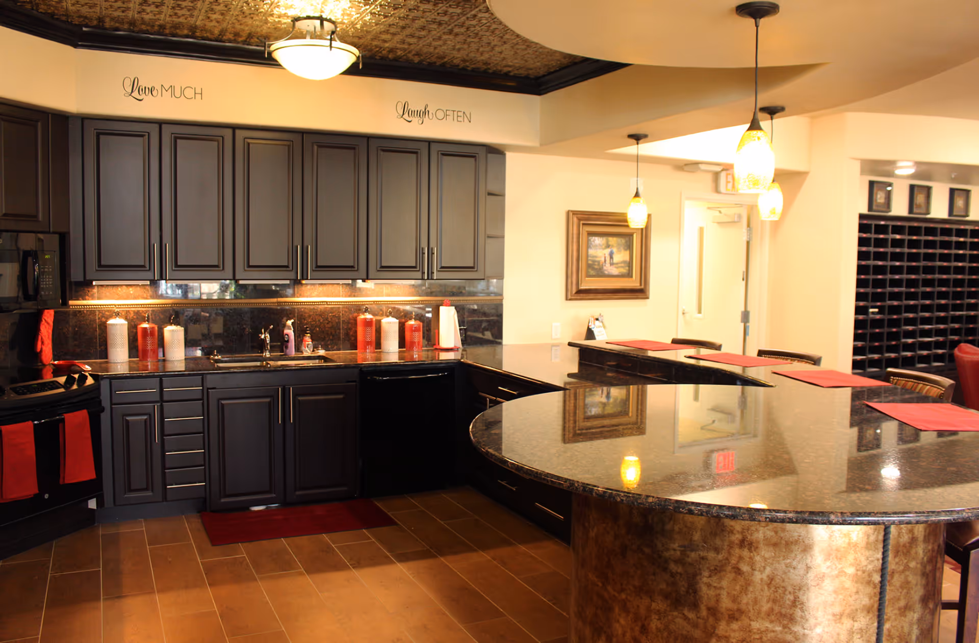 A modern kitchen area with dark wooden cabinets, a black countertop, and a curved island with a polished surface. The kitchen features a microwave, stove, sink, and several decorative canisters. There are pendant lights hanging above the island, and red accents including towels and placemats. On the wall above the cabinets, the phrases 'Love Much' and 'Laugh Often' are displayed. A framed painting and a mail sorting area are visible in the background.