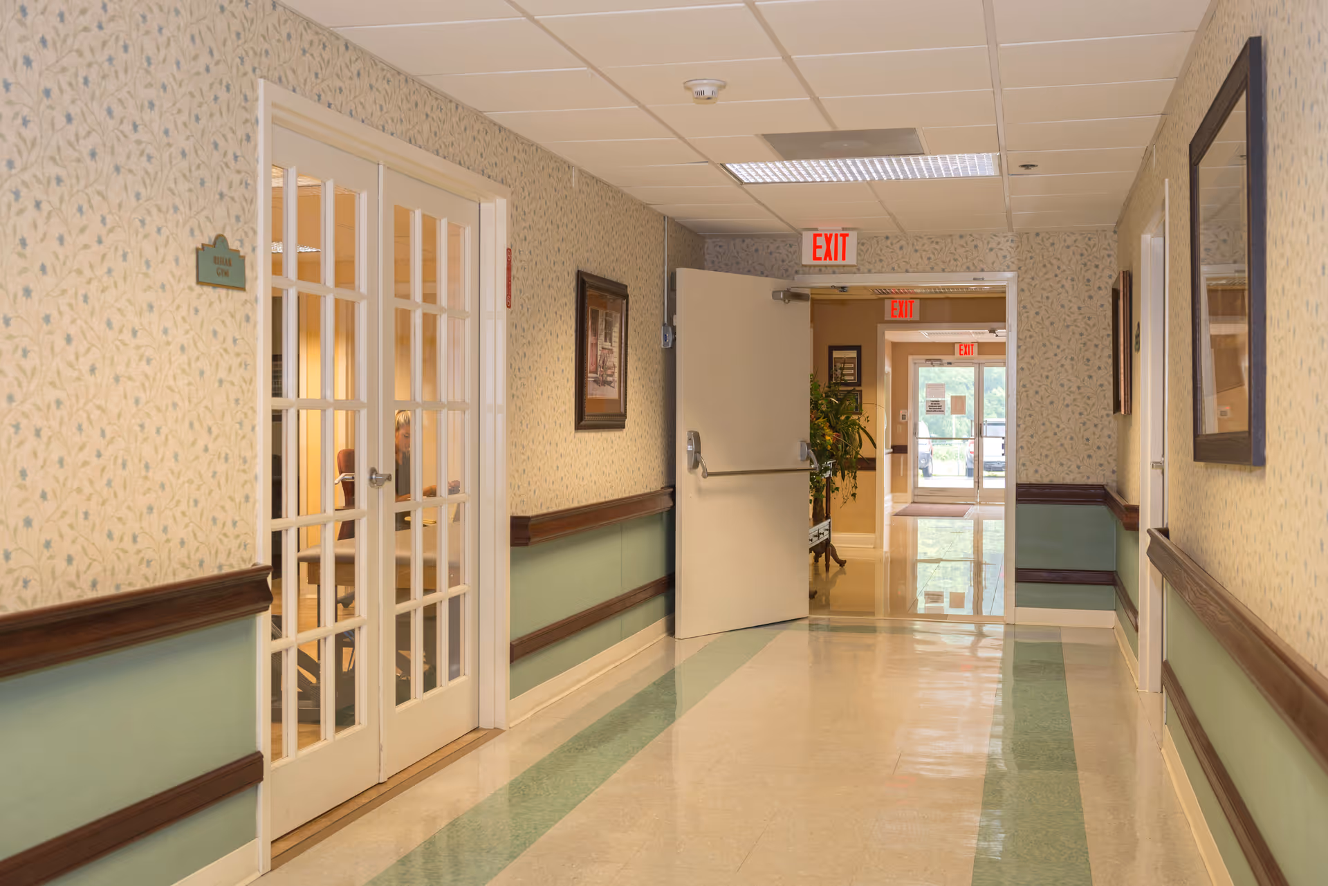 A clean, well-lit hallway in a healthcare facility with patterned wallpaper and green wainscoting. On the left, there are double glass doors leading to a room labeled 'Nursing Gym' where a person is partially visible sitting inside. At the end of the hallway, an open door leads to another corridor with exit signs and a glass door showing an outdoor view.