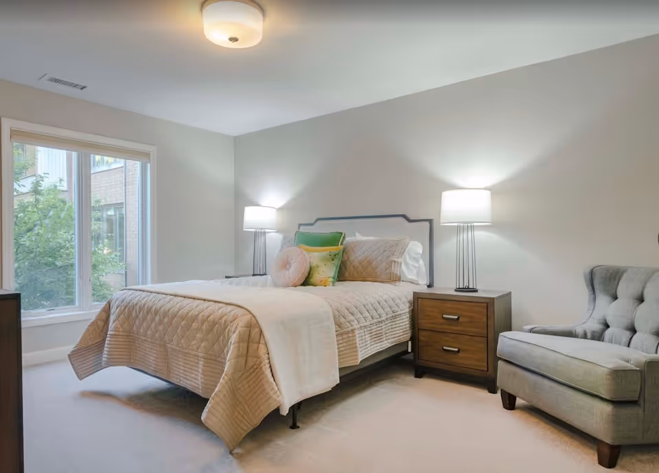 A neatly made bed with a beige quilted bedspread and multiple decorative pillows in a softly lit bedroom. There are two bedside tables with matching lamps on either side of the bed. A gray upholstered armchair is positioned to the right of the bed. A large window on the left side lets in natural light and shows some greenery outside.