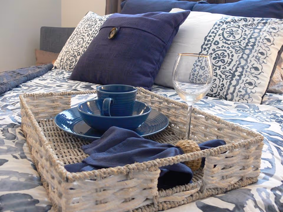 Woven serving tray holding blue dishes, a napkin and a wine glass on a patterned bed with decorative pillows.