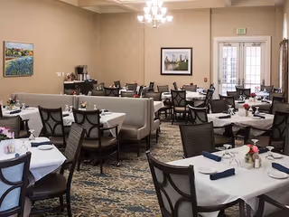 Dining room with multiple tables set with white tablecloths, chairs, and place settings in a communal senior living facility.
