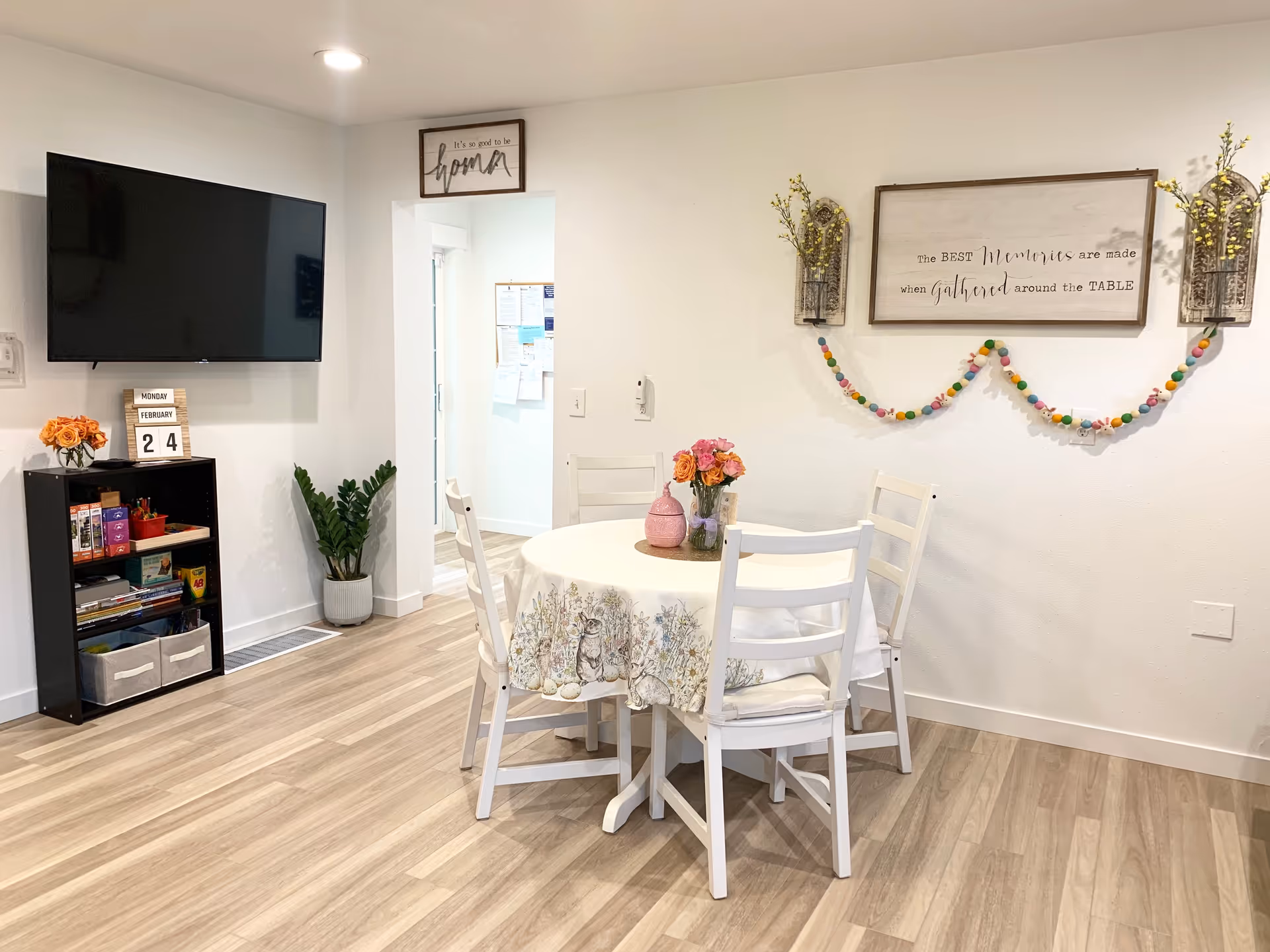 A bright and cozy dining area with a round table covered with a floral tablecloth and four white chairs. On the table, there are two vases with flowers. The wall behind the table has a decorative sign that reads 'The BEST Memories are made when gathered around the TABLE' with two wall-mounted flower holders and a colorful garland. To the left, there is a black shelving unit with books and storage bins, a potted plant, and a wall-mounted flat-screen TV. Above the doorway, there is a small sign that says 'It's so good to be home.' The floor is light wood, and the walls are white.