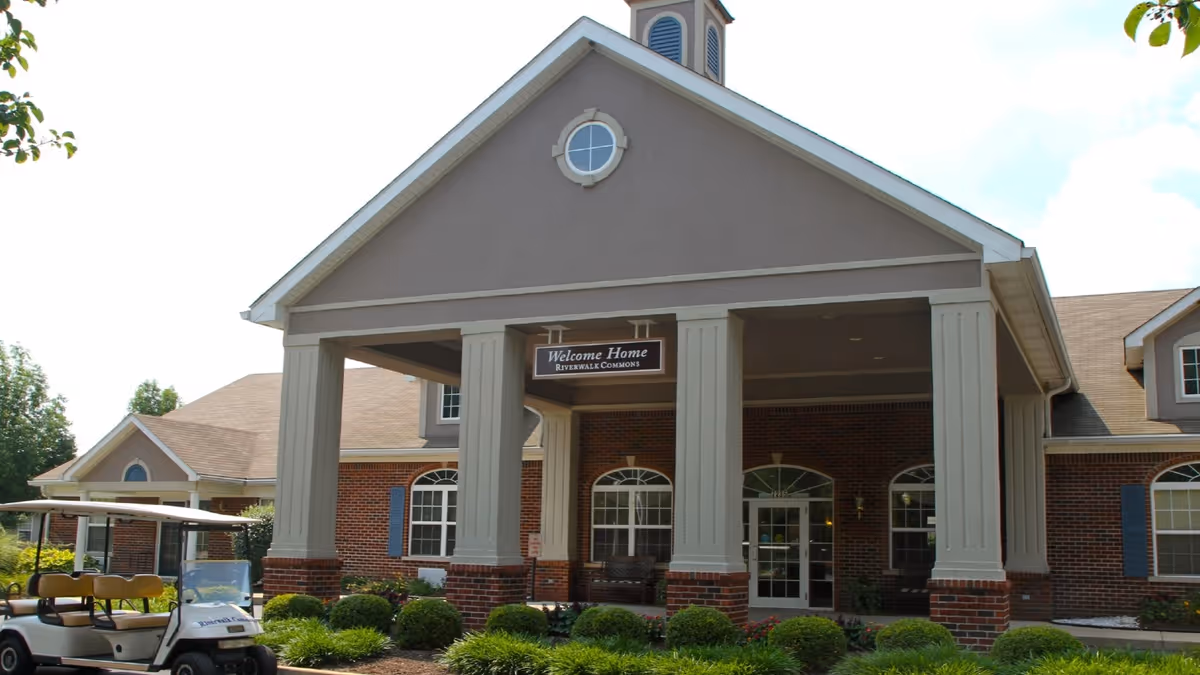 Exterior view of a senior living facility entrance with a covered porch supported by large columns. The building has brick walls, multiple windows with blue shutters, and a sign hanging from the porch roof that reads 'Welcome Home Riverwalk Commons'. There is a golf cart parked to the left and neatly trimmed bushes and greenery in front of the building.