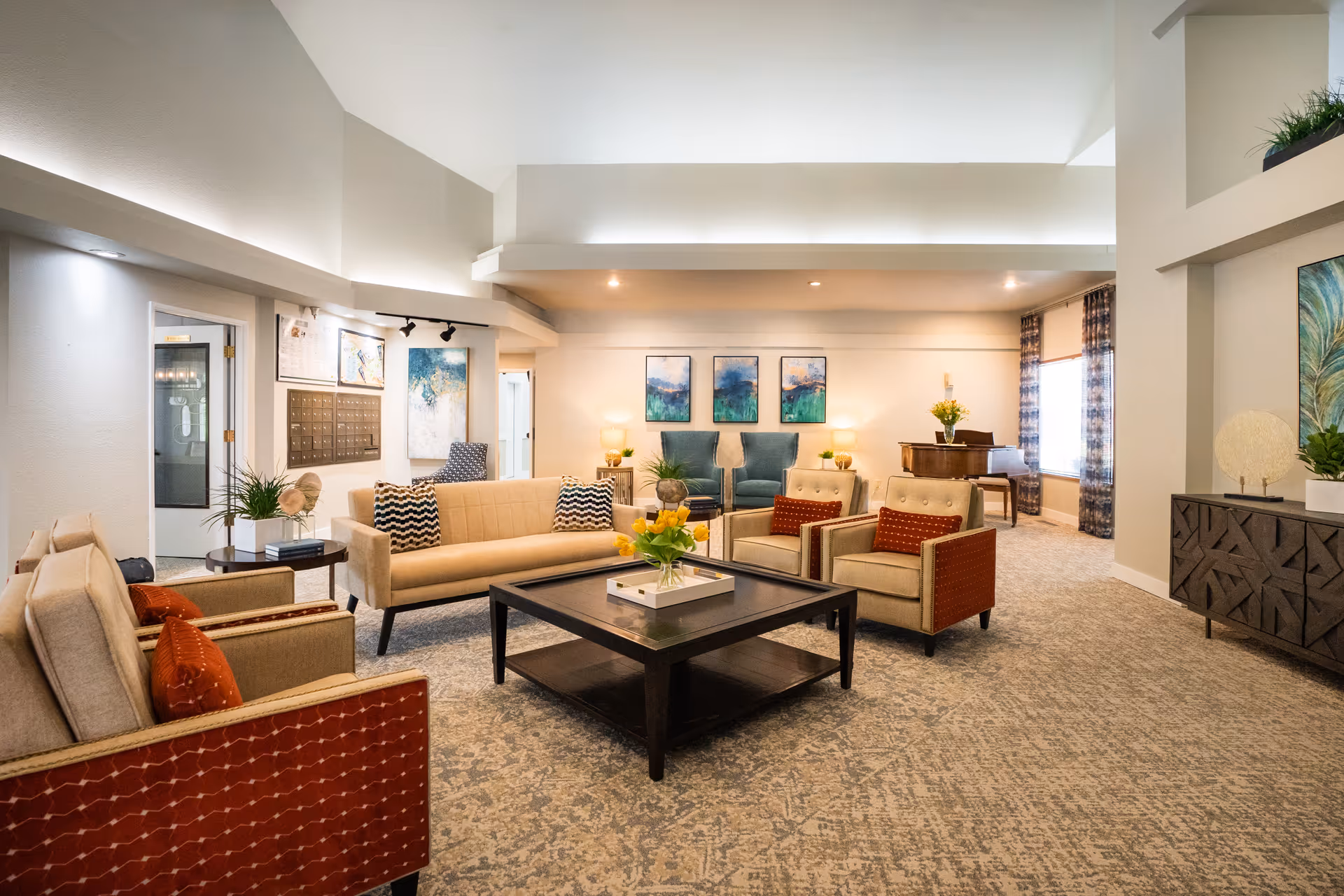 A spacious and well-lit living room area with beige and red armchairs, a beige sofa with patterned pillows, a dark wooden coffee table with a flower arrangement, and two blue armchairs against the far wall. The room features carpeted flooring, modern artwork on the walls, a piano in the corner, and decorative plants and lamps throughout.