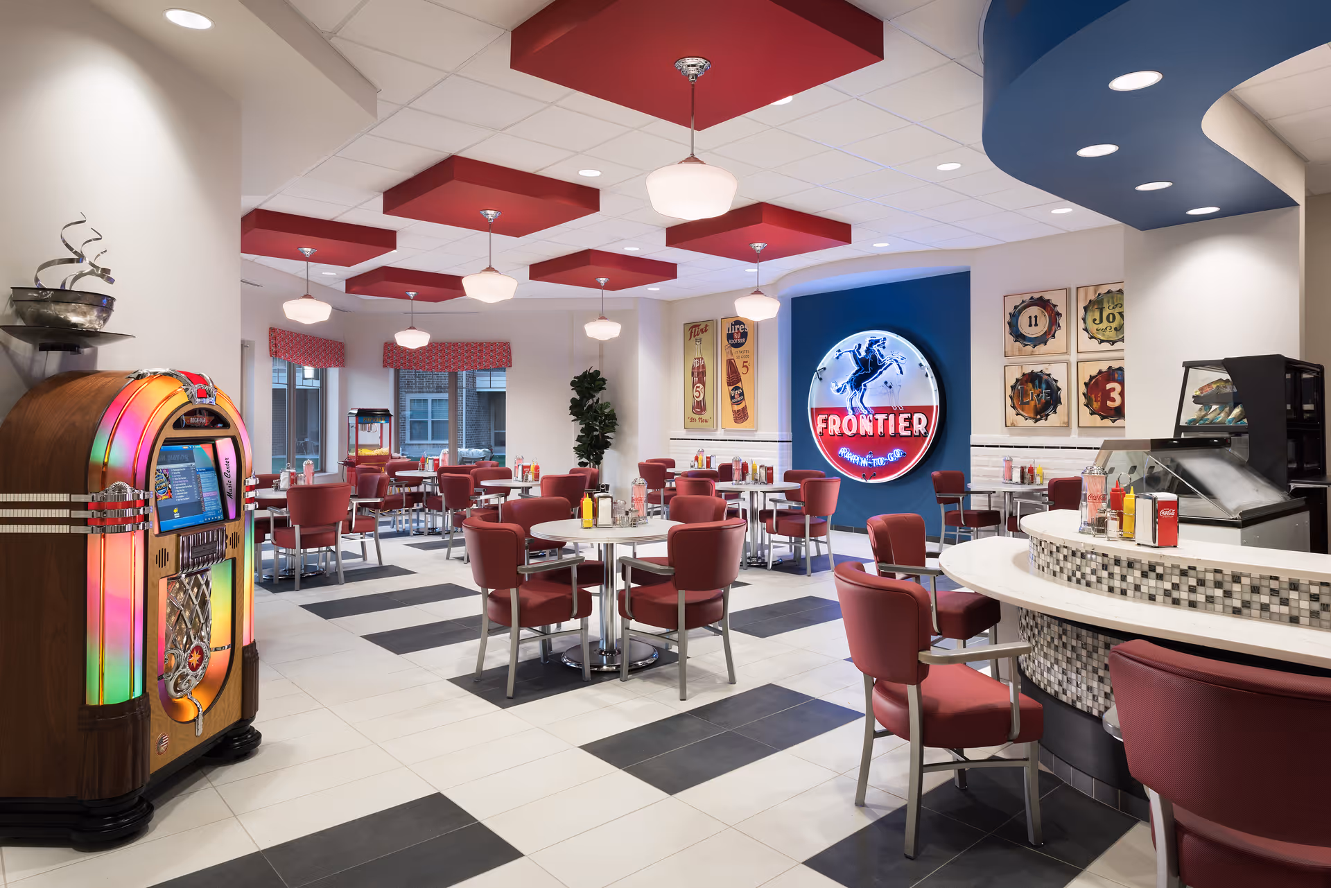 Bright retro-style dining room with red chairs and round tables, a jukebox, counter seating, and a illuminated 'Frontier' sign on the wall.