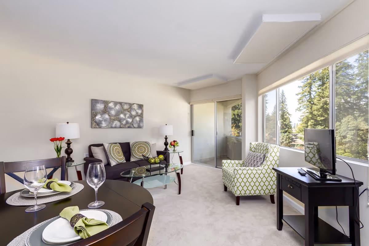 A modern living room featuring a dining table set with plates and glasses, a comfortable sofa with decorative pillows, a glass coffee table, and a green patterned armchair. Large windows allow natural light and showcase trees outside.