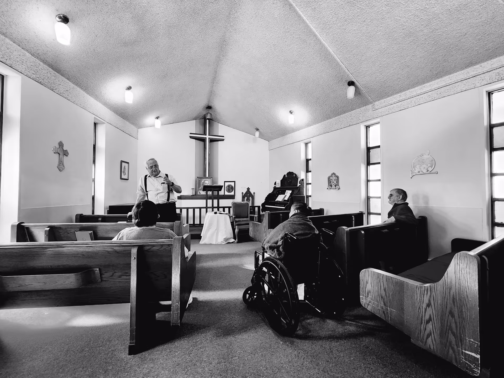 Black and white photo of a small chapel interior with wooden pews and a cross on the wall behind a lectern. Four elderly people are seated in the pews, one in a wheelchair, and an elderly man is standing near the front speaking or gesturing. The room has several windows and wall decorations.