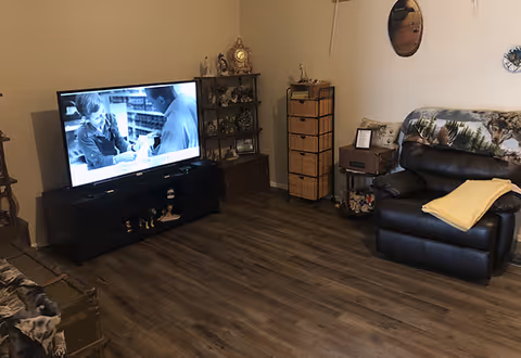 Cozy living room with a television on a stand, a leather recliner with a throw, shelving and side tables, and wood-look flooring.