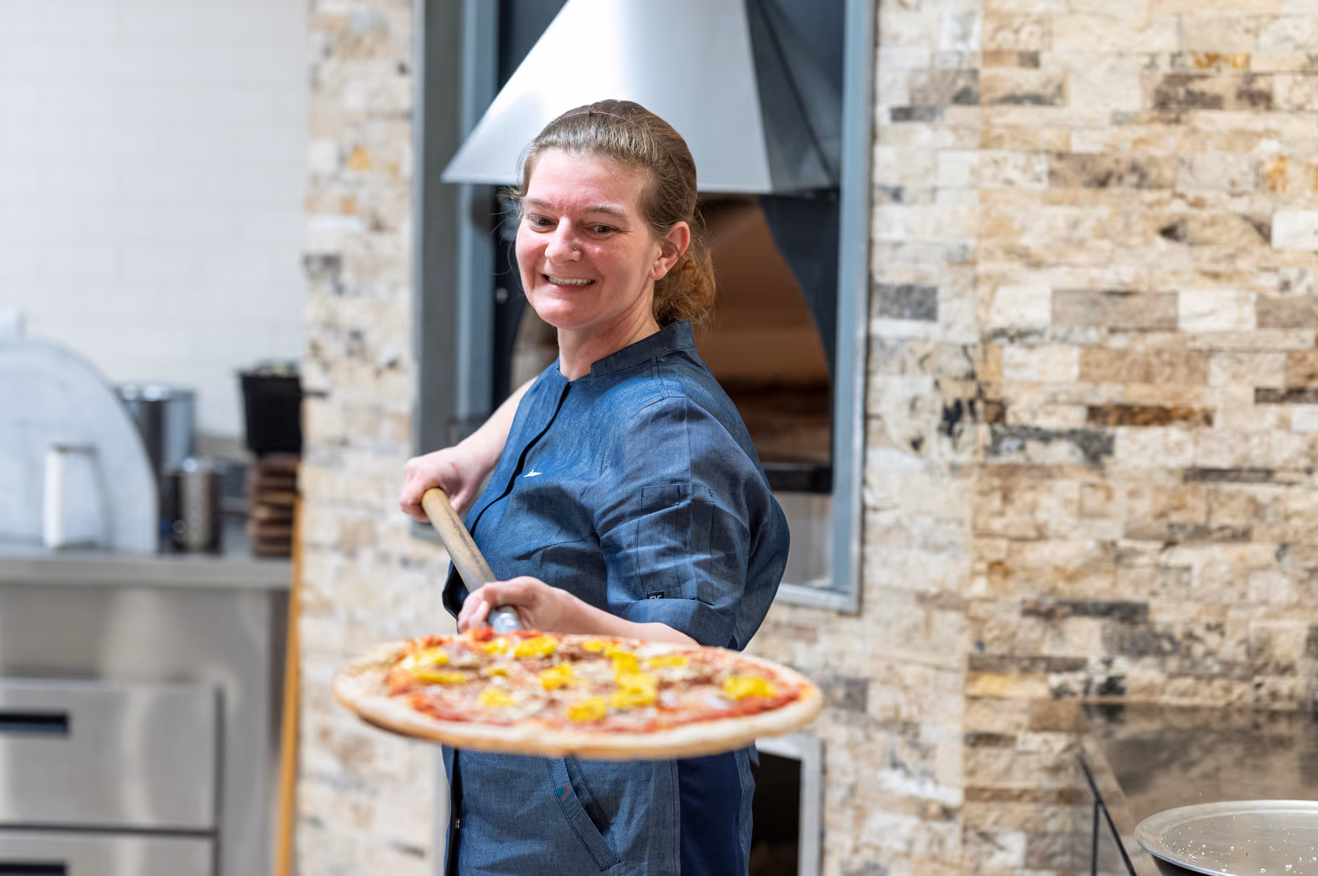A woman in a blue chef's jacket is holding a wooden pizza peel with a freshly baked pizza topped with cheese and other ingredients in a kitchen with a stone wall and pizza oven in the background.