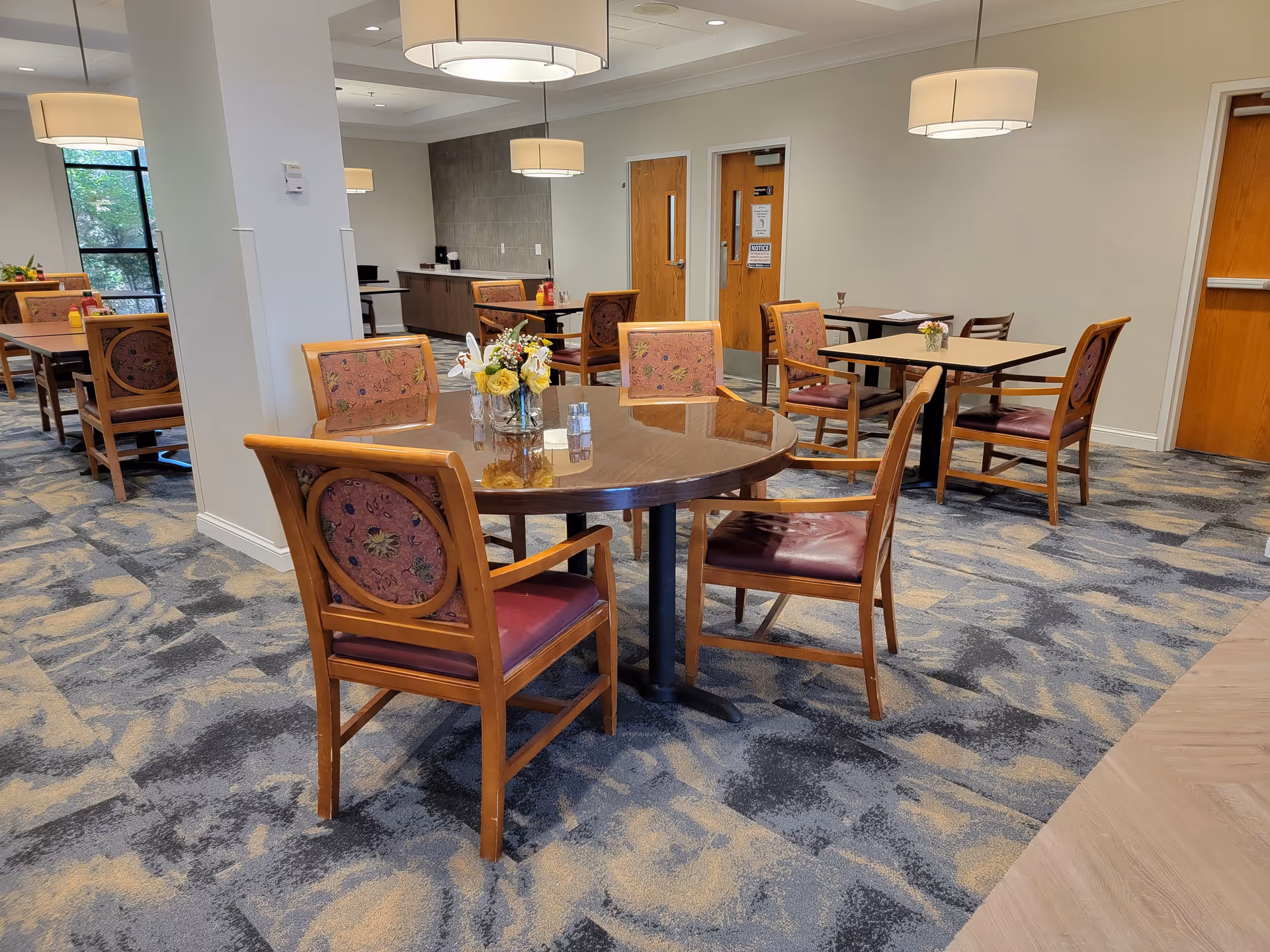 Interior view of a dining area in an assisted living residence featuring multiple wooden tables with floral centerpieces and chairs with patterned upholstery. The room has carpeted flooring with a blue and beige pattern, beige walls, and modern ceiling lights. There are wooden doors and a large window letting in natural light.
