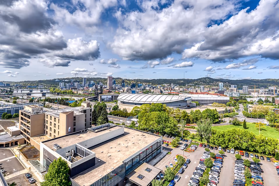 Aerial view of a cityscape featuring a large stadium with a white roof surrounded by trees, buildings, and a parking lot filled with cars under a partly cloudy blue sky.
