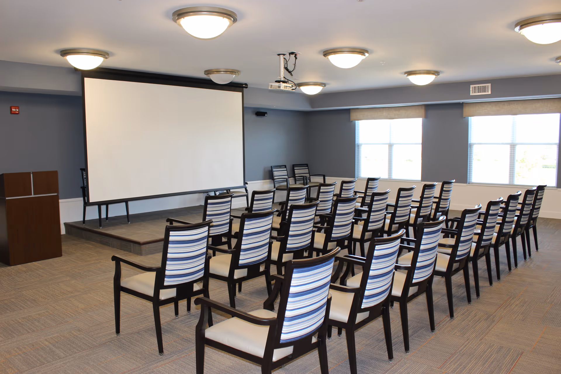 A conference or presentation room with rows of chairs facing a large white projection screen. The chairs have dark wooden frames with striped blue and white backs and beige seats. There is a small stage with a podium on the left side and large windows on the right side letting in natural light. Ceiling lights illuminate the room.