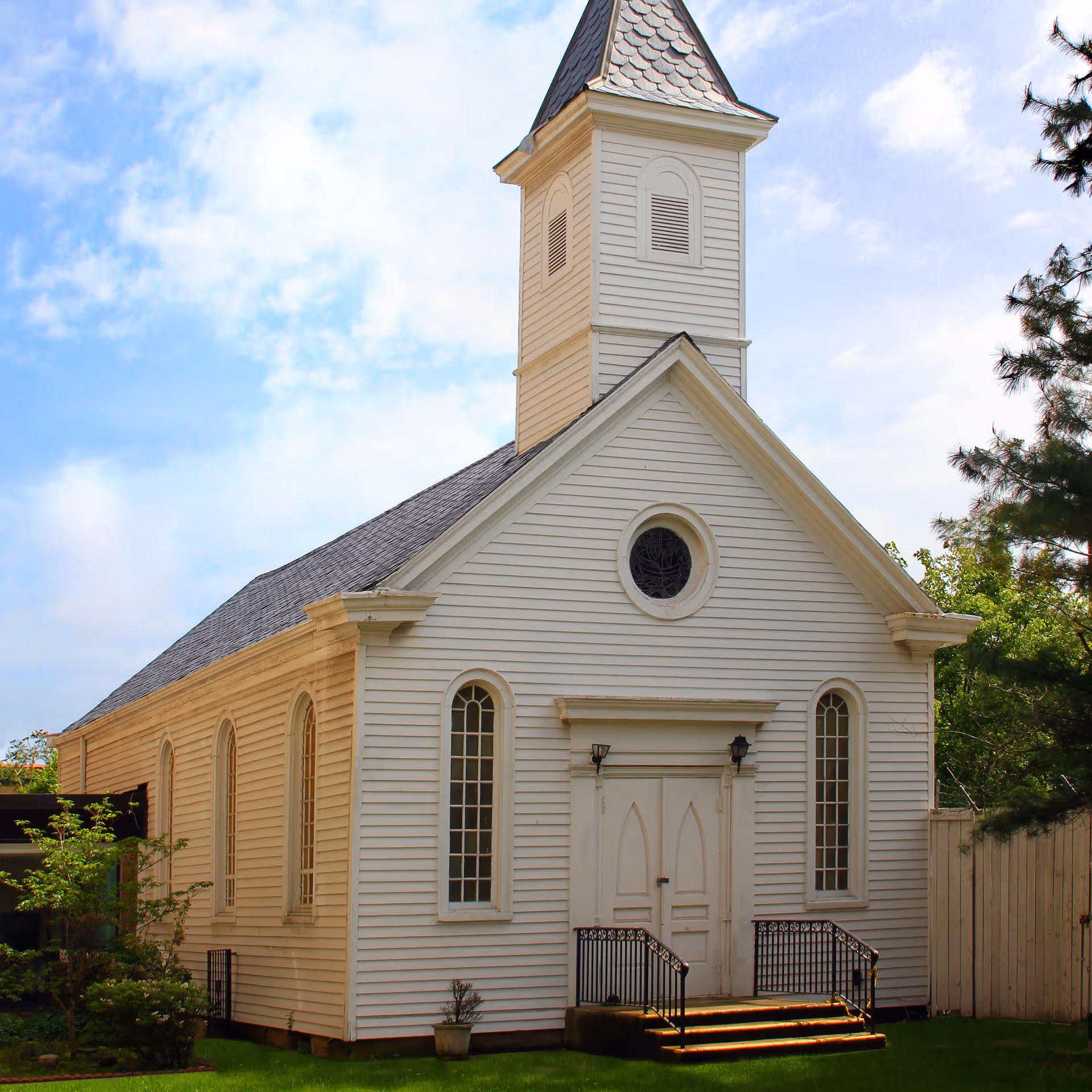 White wooden church building with a tall steeple, arched windows, and double front doors, surrounded by green grass and trees under a partly cloudy sky.