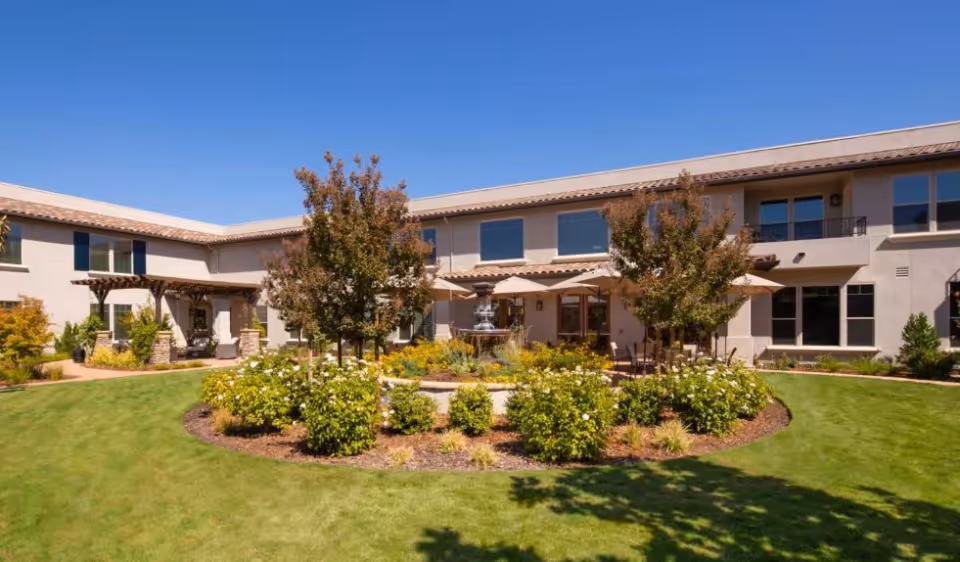 A sunny outdoor courtyard at Oakmont of Fair Oaks featuring a circular garden with green bushes and small trees surrounding a central water fountain. The courtyard is bordered by a two-story building with large windows and a tiled roof under a clear blue sky.