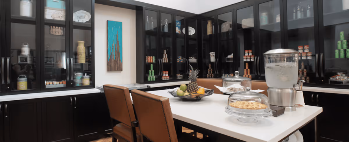 A well-organized pantry or snack area with dark wood cabinets featuring glass doors filled with canned goods and jars. In the center, there is a white countertop island with brown leather chairs around it. On the island, there is a fruit bowl with a pineapple and other fruits, a glass container with a pie, a water dispenser, and some cups.