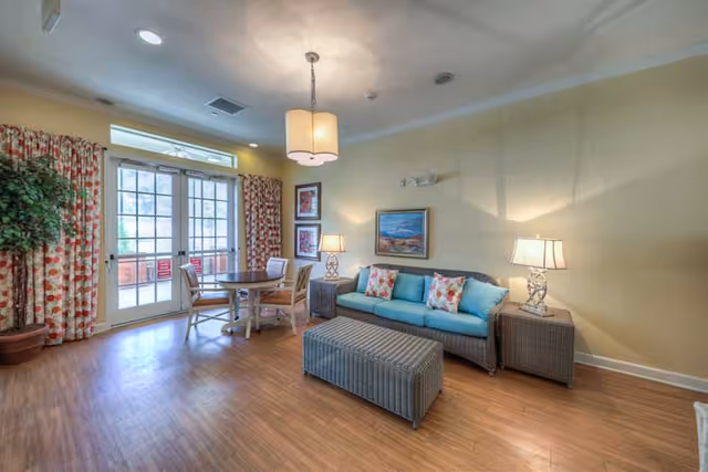 A cozy living room area with a wicker sofa featuring blue cushions and floral pillows, a matching ottoman, two side tables with lamps, a round wooden table with four chairs, floral curtains on the windows and glass doors leading outside, and framed artwork on the light yellow walls.