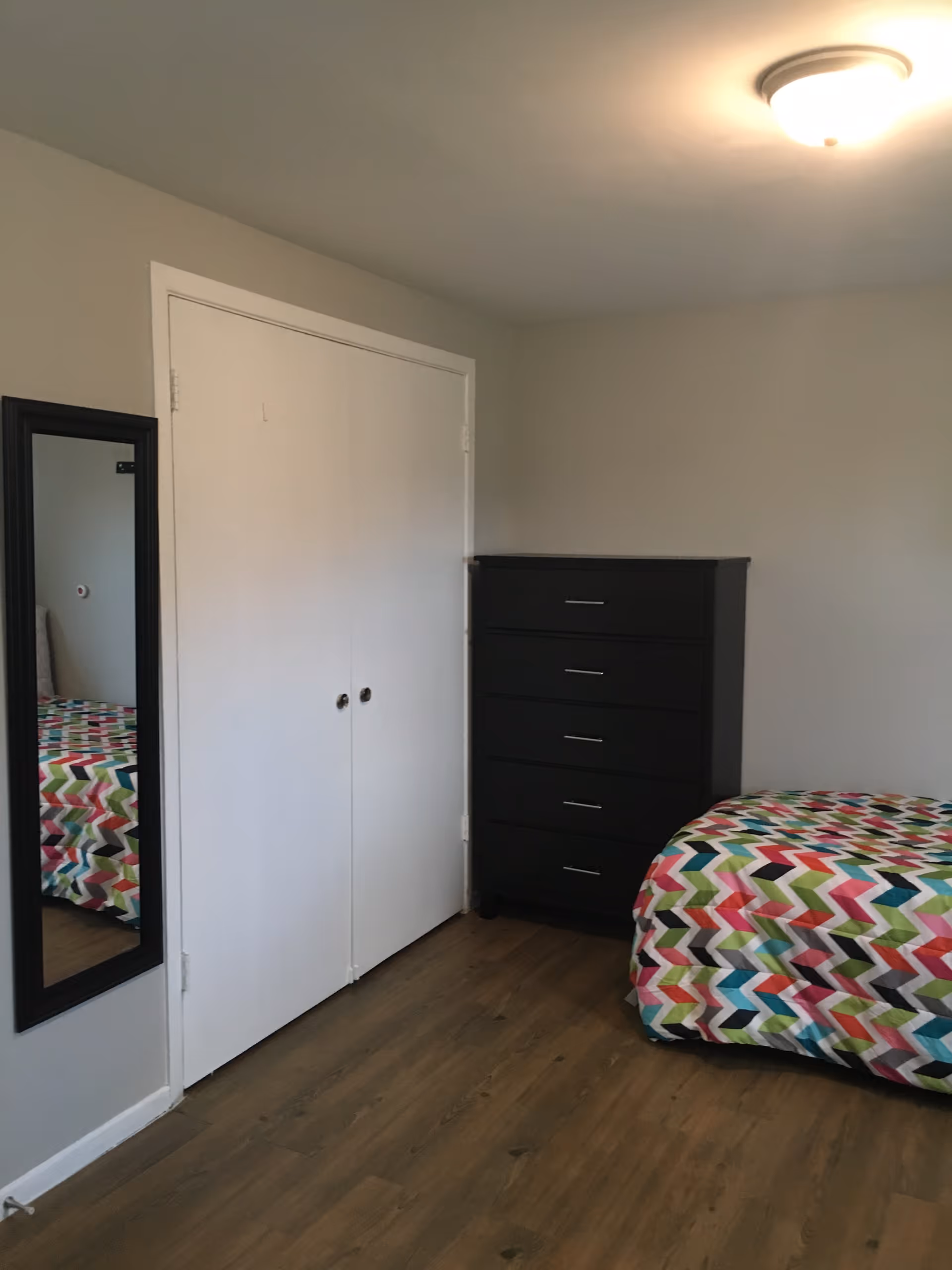 A bedroom with a colorful zigzag patterned bedspread on a bed, a black chest of drawers, a white double-door closet, a full-length mirror with a black frame mounted on the wall, and wooden flooring.