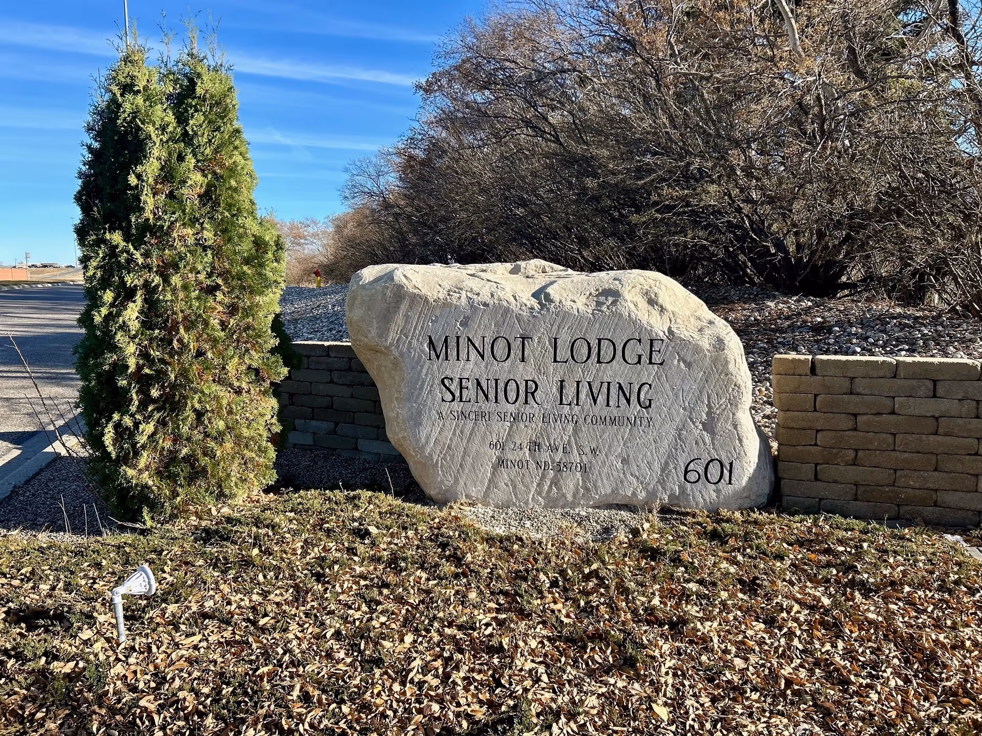 A large rock sign at the entrance reads Minot Lodge Senior Living, with surrounding landscaping.