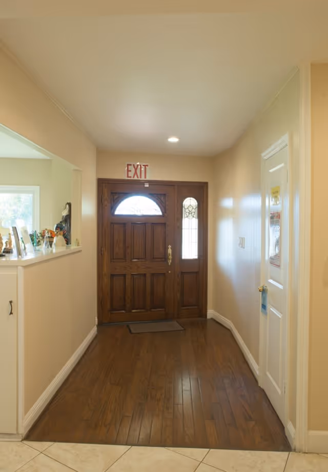 Interior hallway with wooden floor leading to a wooden front door with decorative glass panels and an illuminated exit sign above it. To the right is a white door with notices attached, and to the left is a half wall with various decorative items on top.