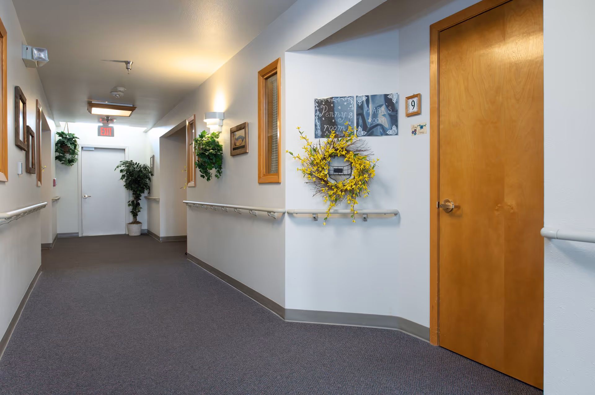 A clean, well-lit hallway in a senior living facility with beige walls, carpeted floor, wooden doors, handrails along the walls, decorative plants, and framed pictures. There is a yellow wreath hanging on the wall near a door labeled 'Penny's room' and 'Mark's room'.