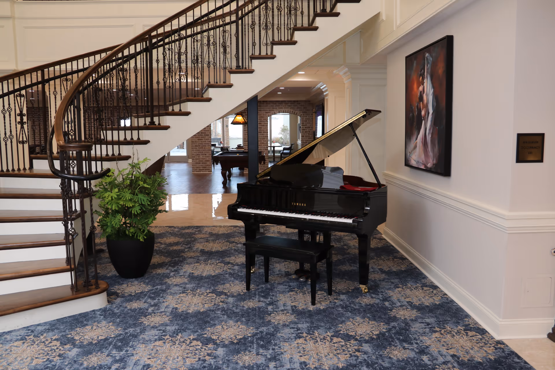 A grand piano sits beneath a curved staircase in a carpeted interior lobby with artwork and a potted plant.