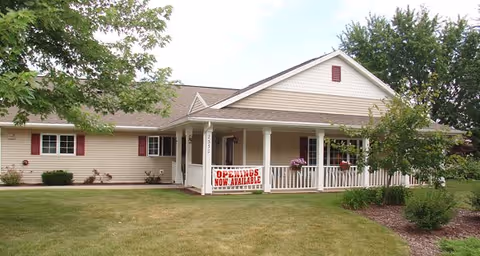 Single-story beige building with white trim and a covered front porch. There is a sign on the porch railing that reads 'OPENINGS NOW AVAILABLE'. The building is surrounded by green grass, trees, and shrubs under a partly cloudy sky.