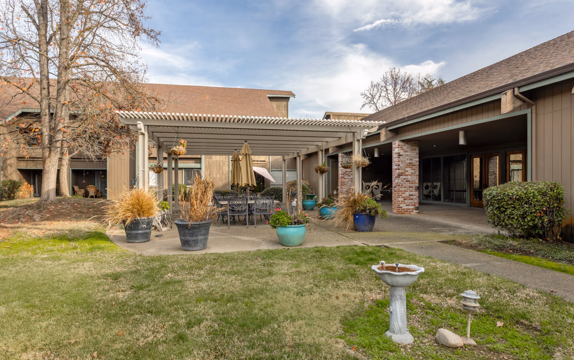 Outdoor courtyard featuring a pergola-covered patio with a dining table, potted plants, and surrounding senior living building.
