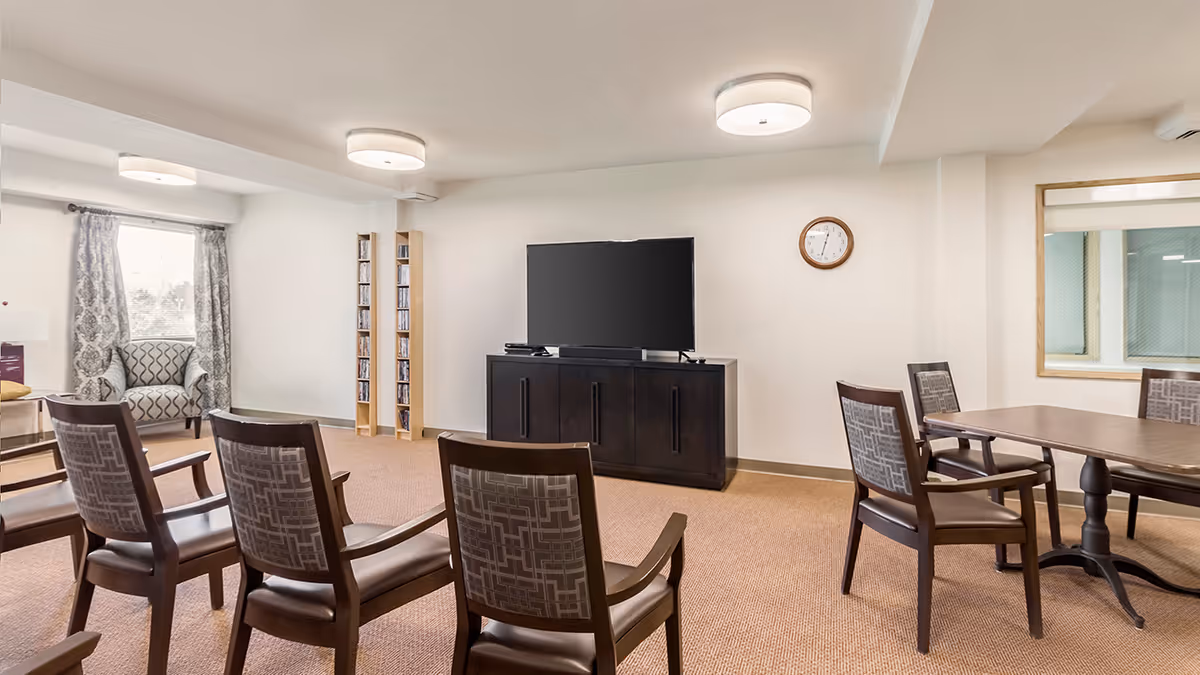 A common area in a retirement community with several wooden chairs arranged facing a large flat-screen TV on a dark wooden cabinet. There is a round clock on the wall, a window with patterned curtains, a patterned armchair, and a table with chairs on the right side. The room has beige carpet and white walls with ceiling lights.
