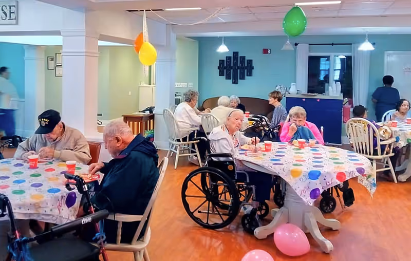 Seniors seated at decorated tables and in wheelchairs socializing in a communal dining/activity room with balloons.