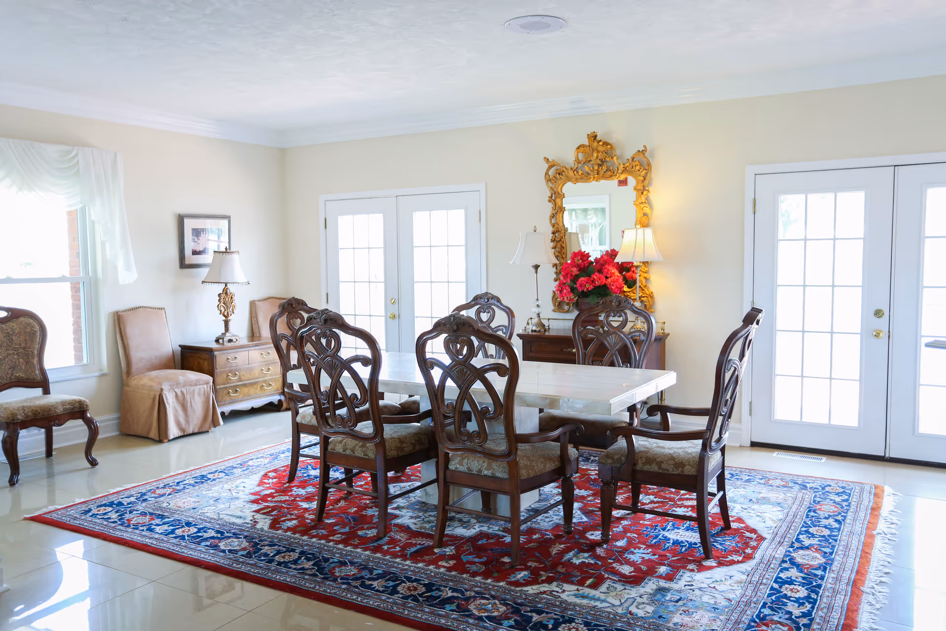 A bright dining room with a large rectangular table surrounded by eight ornate wooden chairs with upholstered seats. The floor is tiled and covered partially by a large, colorful patterned rug. There are two sets of French doors letting in natural light, a wooden side table with a lamp and a framed picture on the wall, and a decorative gold-framed mirror above a small console table with a lamp and a vase of red flowers.
