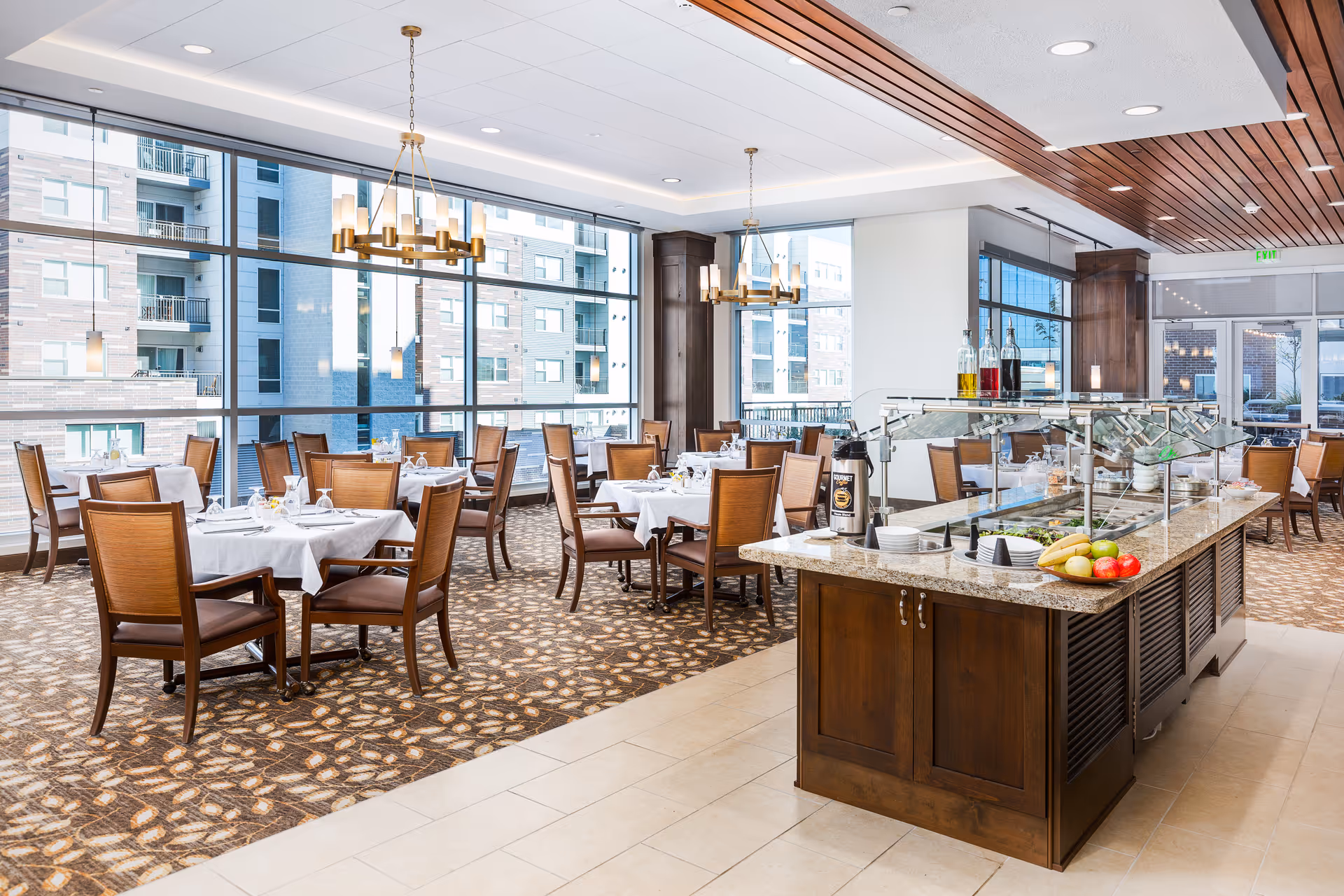 Bright dining room with large windows showing an outside view of nearby buildings. The room features multiple tables covered with white tablecloths, each set with glassware and napkins. There are wooden chairs around the tables and two modern chandeliers hanging from the ceiling. In the foreground, there is a buffet counter with a granite top, holding plates, bowls, condiments, and fresh fruit.