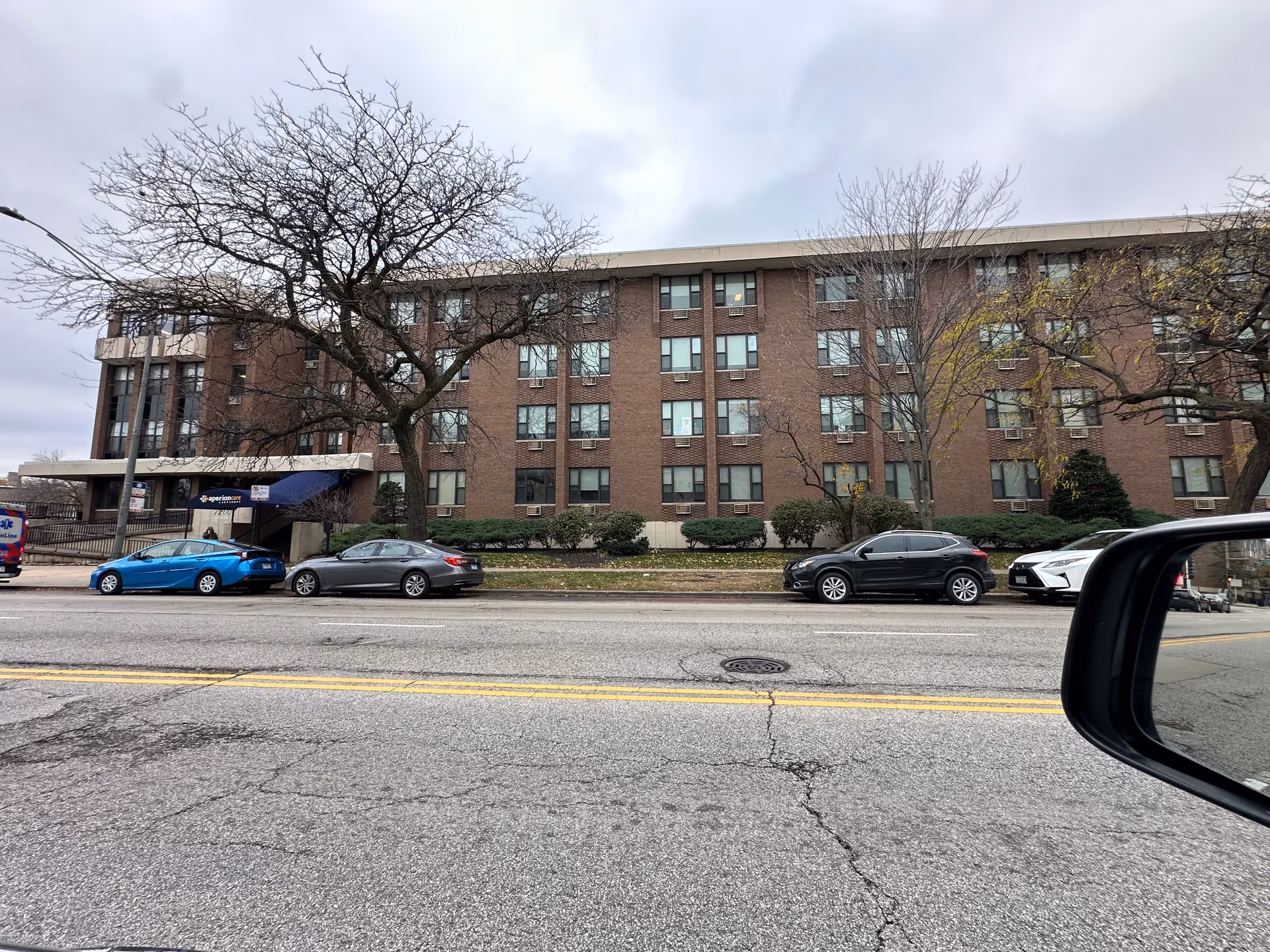 Four-story brick senior living building with a blue entrance canopy, parked cars along the street and bare trees under a cloudy sky.