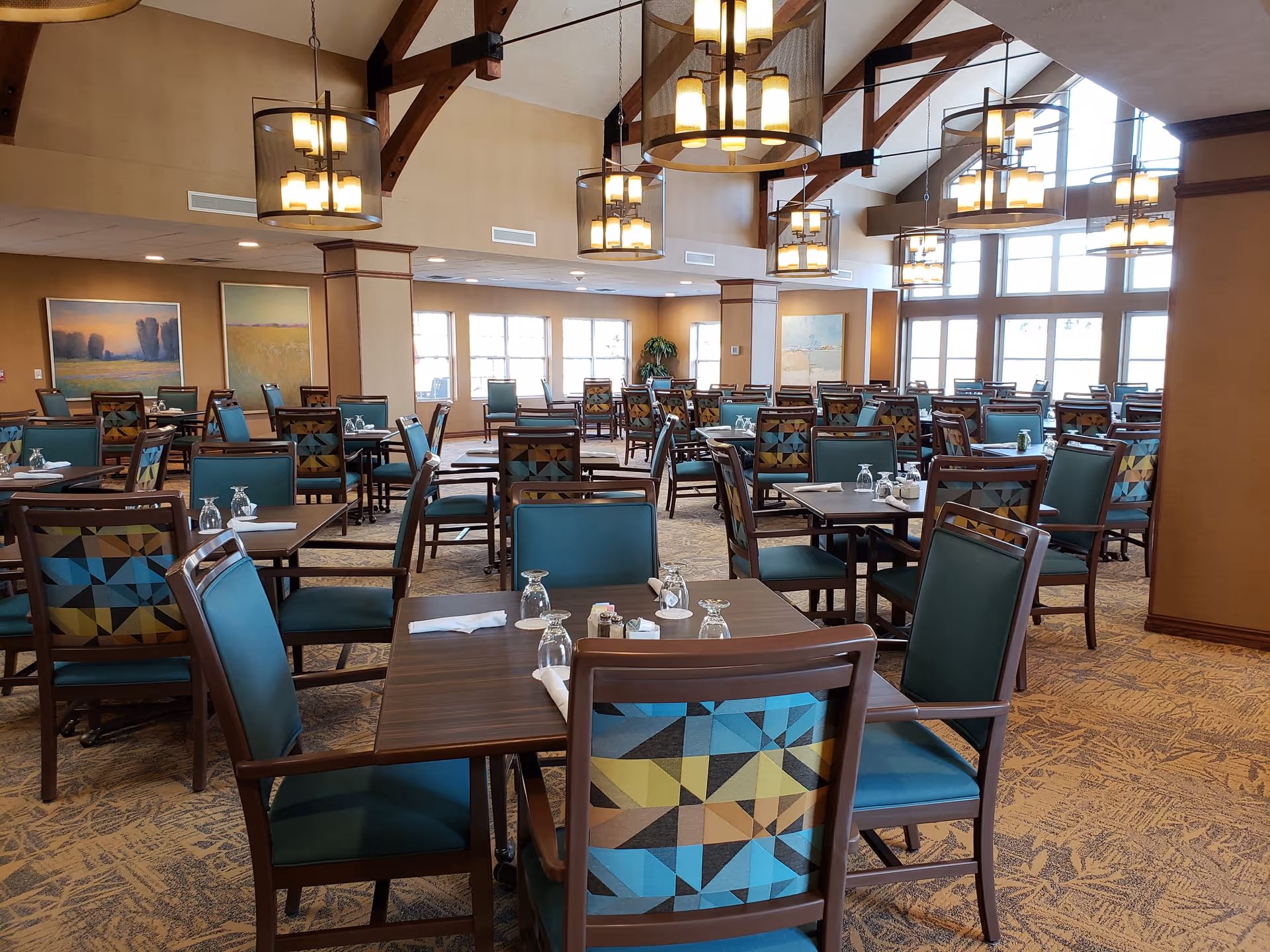A spacious dining room with multiple wooden tables and chairs featuring teal and patterned upholstery. The room has large windows letting in natural light, beige walls, carpeted floors, and several modern hanging light fixtures with exposed wooden beams on the ceiling.