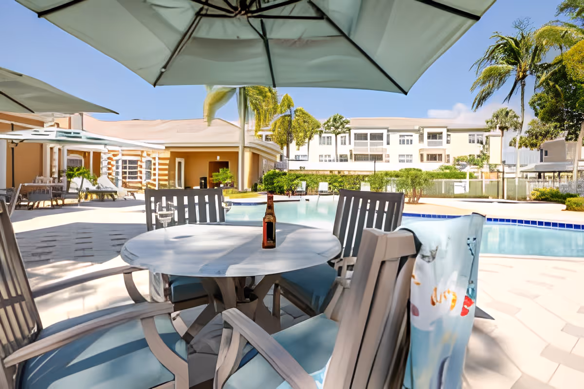Poolside patio with a round table and chairs under umbrellas overlooking a swimming pool and nearby residential buildings.