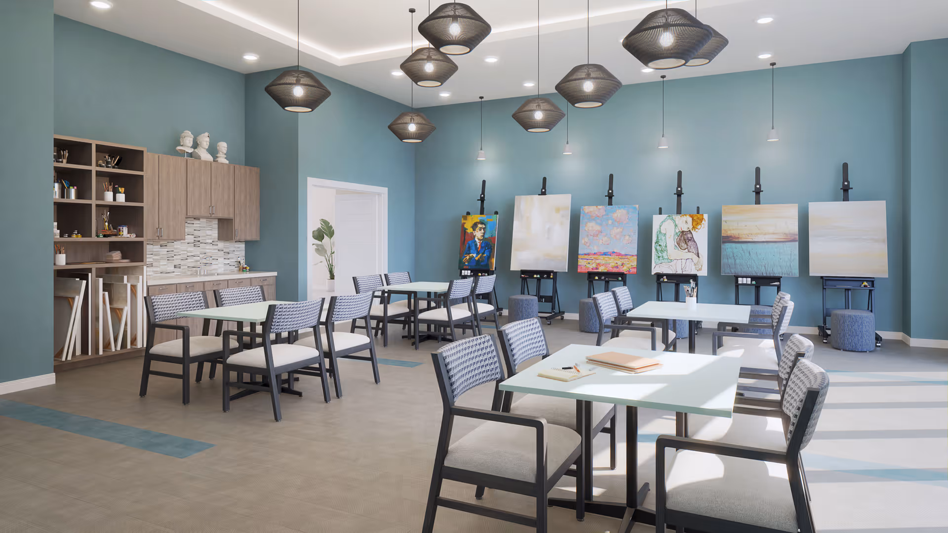 Bright communal activity room with tables and chairs and several easels displaying paintings against teal walls.