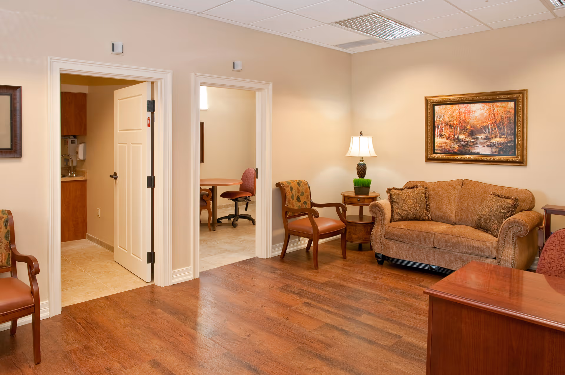 A cozy living room area in a senior living facility with a brown upholstered sofa, two wooden chairs with patterned cushions, a wooden side table with a lamp and a small plant, and a framed painting of an autumn forest scene on the wall. The room has wood flooring and beige walls. Two open doorways lead to adjacent rooms, one showing a kitchenette and the other a small office or meeting room with a round table and office chair.