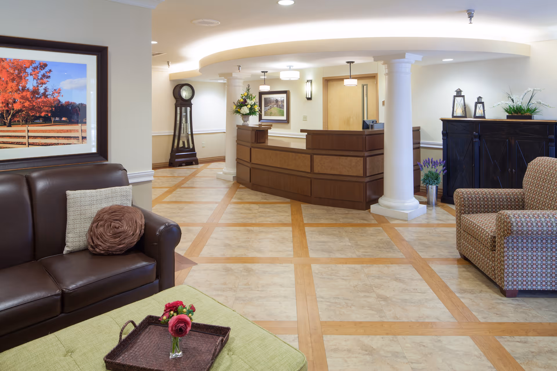 A well-lit reception area in a senior living facility featuring a wooden reception desk with a flower arrangement, a grandfather clock in the corner, a brown leather sofa with decorative pillows, a patterned armchair, a green ottoman with a tray holding a small vase of flowers, and framed artwork on the walls.